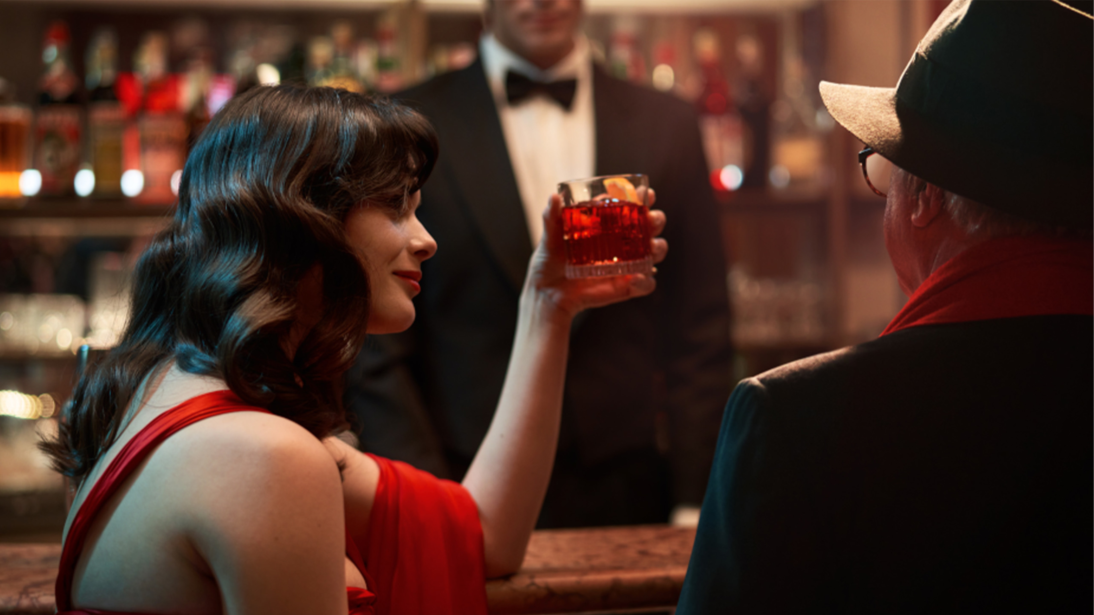 Woman in a red dress sits at a bar with a Campari drink.
