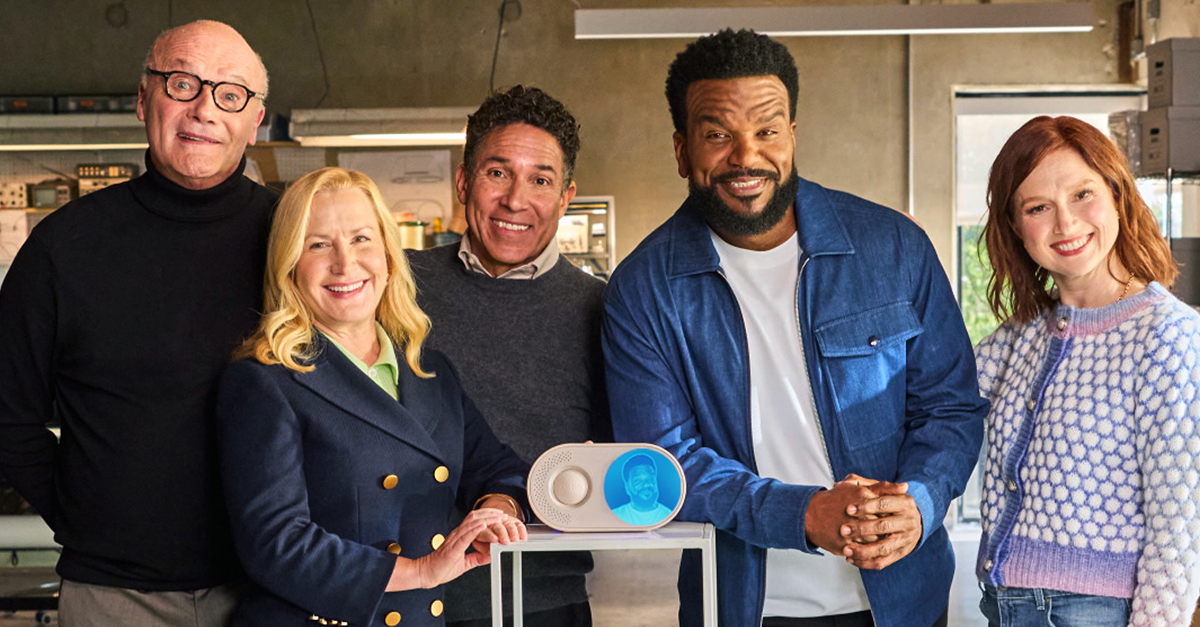 Craig Robinson and cast members gather around a small device showing a blue character.