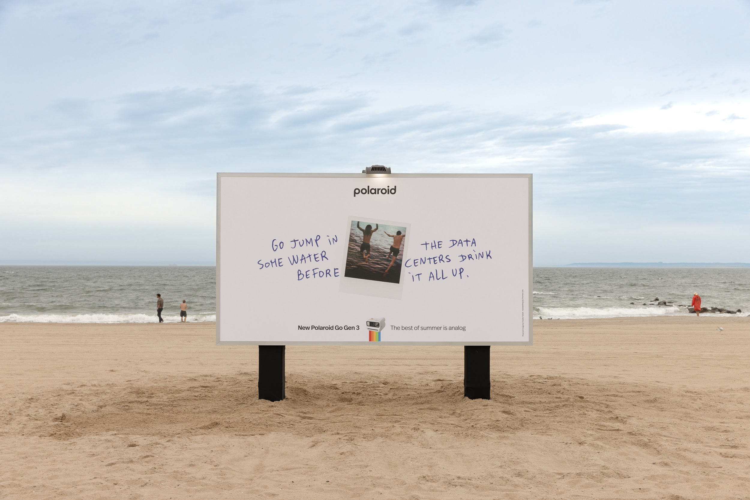 A giant Polaroid billboard rises from Coney Island beach with a line about data centres drinking the water.