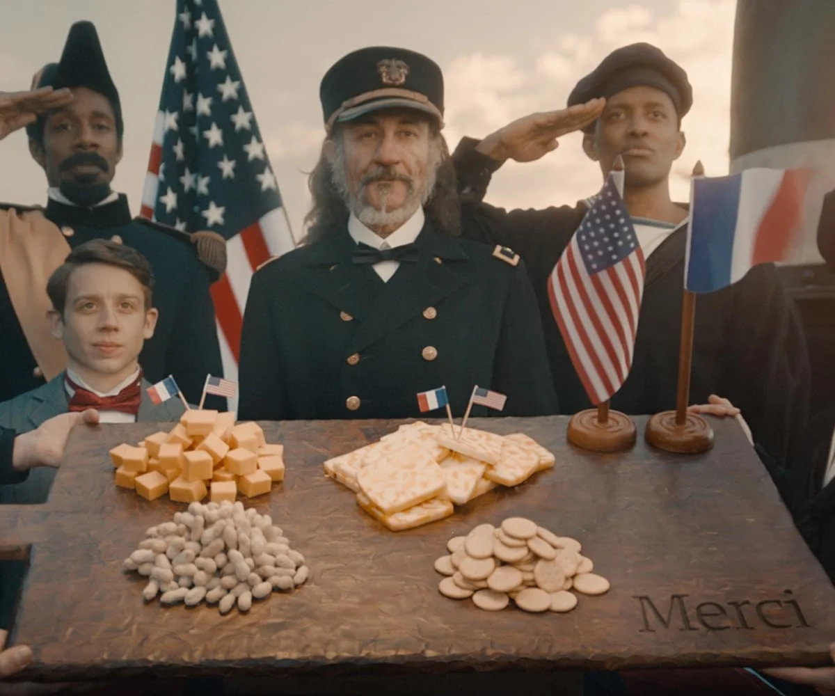 Costumed figures salute behind a large cheese board with American and French flags and the word Merci.