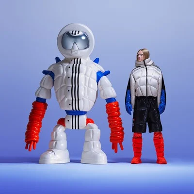 A white, rounded sculptural explorer beside a person in a white puffer jacket, black shorts and red legwear against a blue studio backdrop.